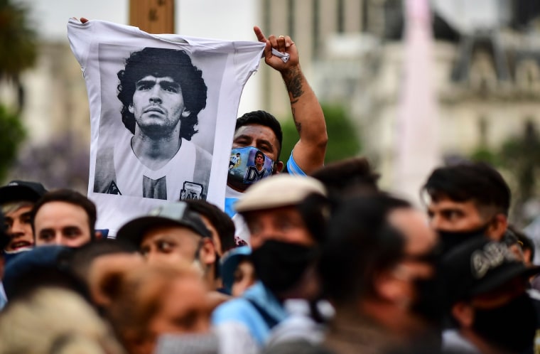 Image: A man waves a t shirt with a picture of Diego Maradona as fans wait to enter the Government House to pay tribute to the late football legend in Buenos Aires