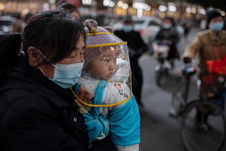 Image: A woman wearing a face mask holds a baby that wears a protective shield during rush hour on a street outside of a shopping mall complex in Wuhan 