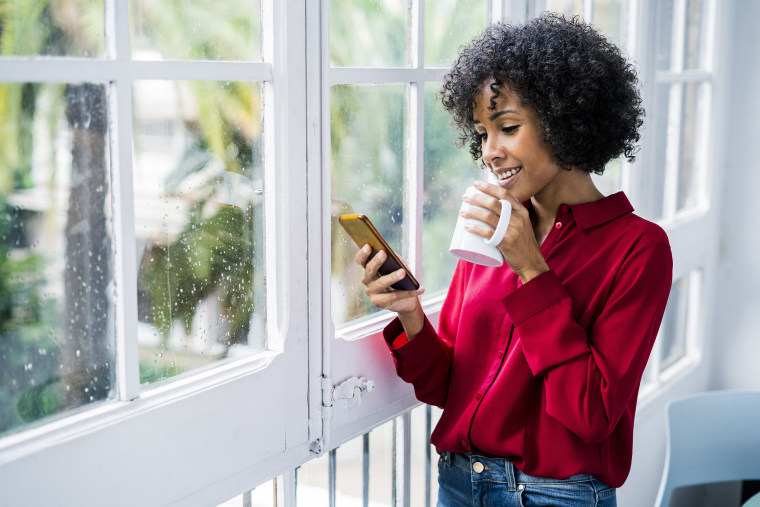 Image: Smiling woman with cup of coffee and cell phone standing at the window at home