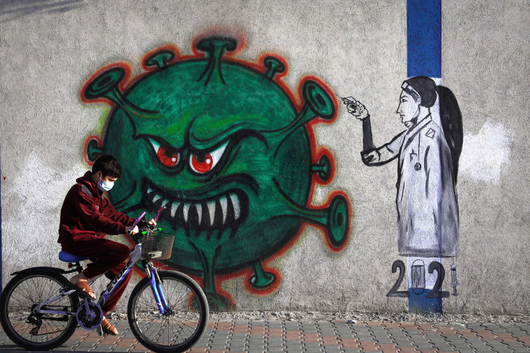 Image: A Palestinian youth wearing a face mask rides his bicycle past a mural painting  of a nurse injecting a vaccine to a Covid-19 virus in Gaza City