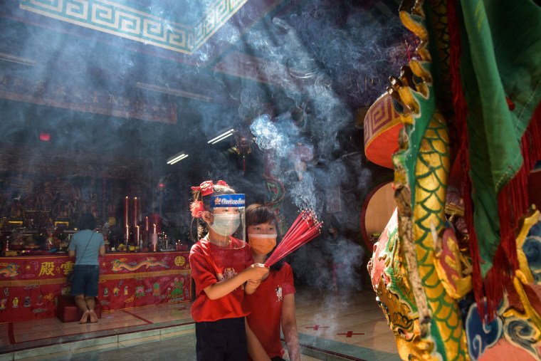 Image: A girl wearing a protective mask and face shield holds incense sticks while praying with her mother