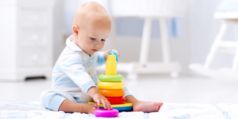 Cute baby playing with colorful rainbow toy pyramid sitting on play mat in white sunny bedroom.