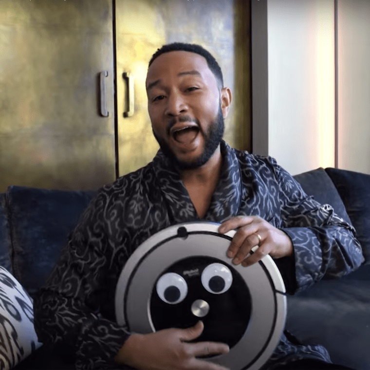John Legend holds a roomba with googly eyes while sitting on a couch in a robe