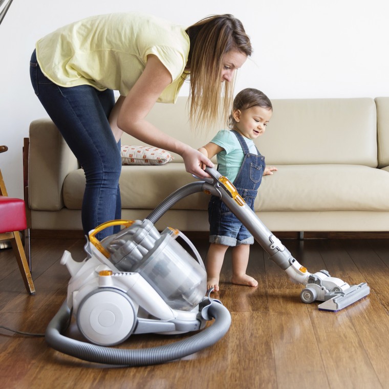 Woman and young boy wearing denim dungarees, standing in front of sofa, hoovering hardwood floor.