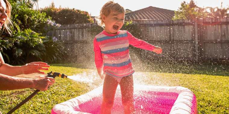 Little girl playing in a pink kiddy pool in the backyard, while her mom sprays her with the house