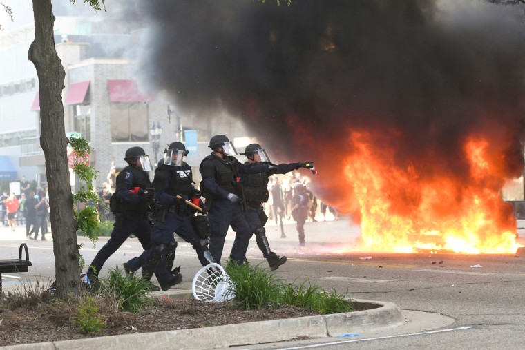 Police move to block off an intersection in downtown Lansing on May 31, 2020, as a car burns in the background.