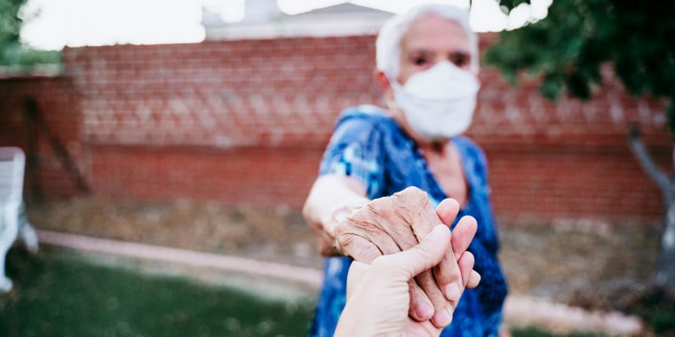 Older woman in backyard wearing an Kn95 mask and holding someones hand. The CDC updated their guidelines on how to shop for face masks and how vaccinated people can socialize. Learn about the updates for face masks and more.