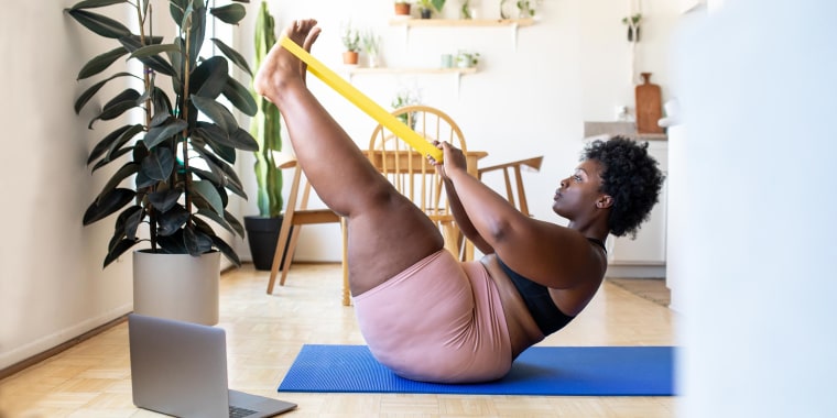 Woman sitting on a blue yoga mat, using a yellow resistance band, to workout on her floor