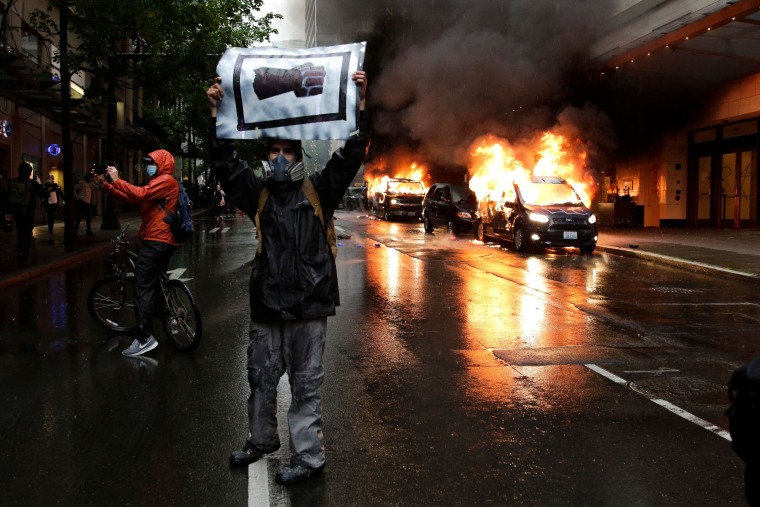 A man holds an image of a fist in front of burning vehicles following demonstrations protesting the death of George Floyd, in Seattle, Wash. on May 30, 2020