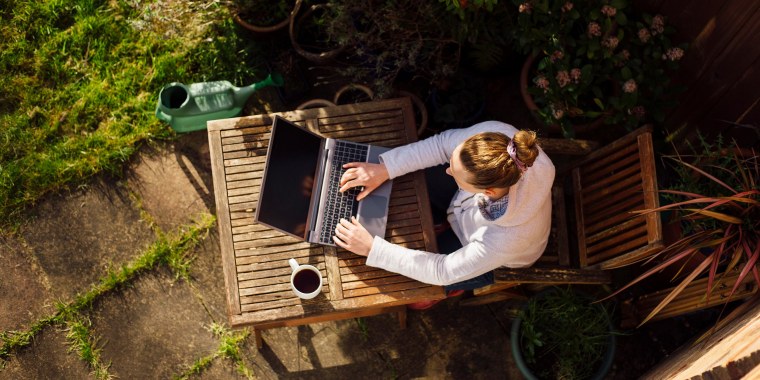 Woman sitting outside with a cup of coffee, surrounded by her garden, working on her laptop. Shop the best outdoor office ideas for working from home outside, including office furniture and accessories such as fans, umbrellas, laptop desks and more.