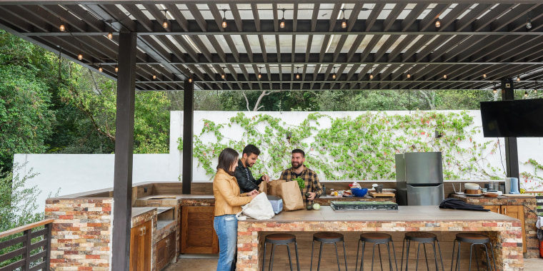 Three people standing in an outdoor backyard kitchen, covered by an overhang, with groceries