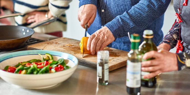 Man cutting a pepper on a wooden chopping board, surrounded by a salad and some olive oil