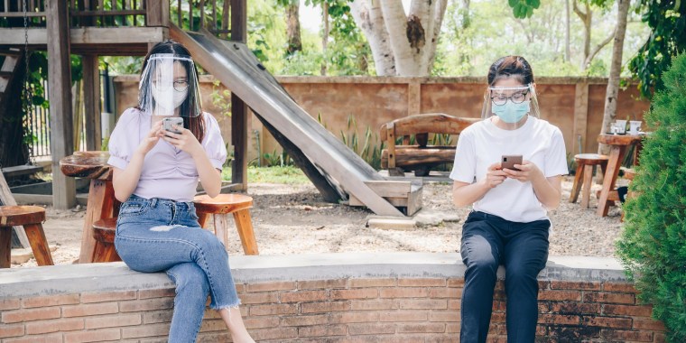 Two Girls 6 feet apart, sitting on a railing at the park wearing face masks and protective eye shields. Shop the best expert-recommended coronavirus eye protection to wear during the pandemic like glasses, goggles and face shields.