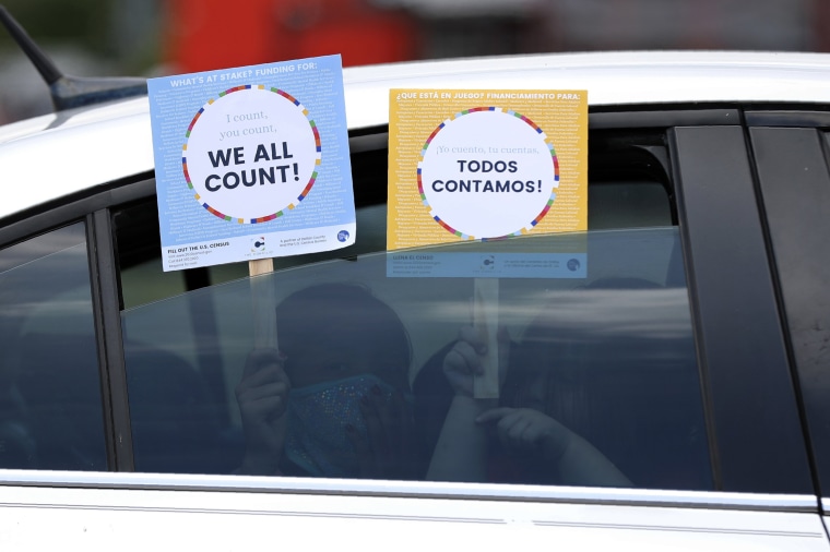 Image: Two young children hold signs through the car window that make reference to the 2020 U.S. Census as they wait in the car with their family at an outreach event in Dallas