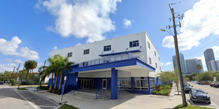 A two-story concrete building with blue balconies sits on a street corner in Miami against a blue sky