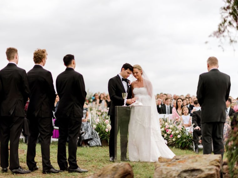 A bride and groom stand side by side in front of a crowd of family and friends outside under an overcast sky