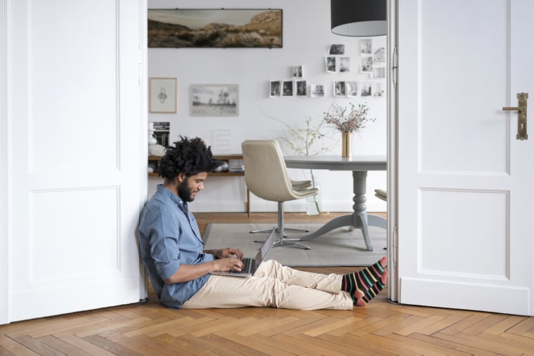 Man at home sitting on floor working with laptop in door frame. Here are the best work from home pants for men in 2021. Check out comfortable pants for men from brands like Everlane, Outdoor Voices, J.Crew and more.