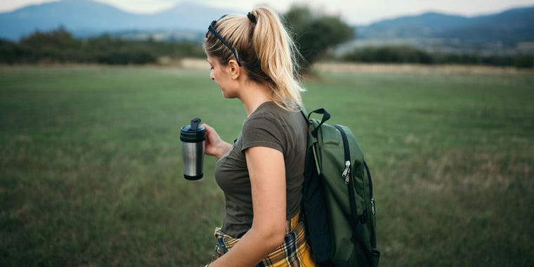 Woman is hiking in nature, with a backpack and holding a travel mug. The best travel mugs of 2021 include reusable cups, travel coffee mugs, ceramic travel mugs and stainless steel travel mugs from Ember, Contigo, YETI and more.
