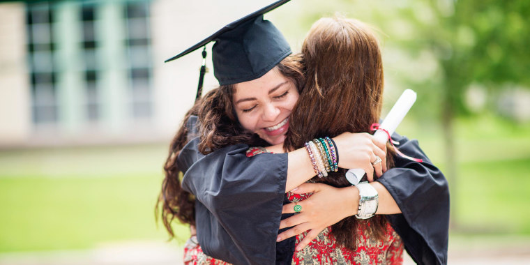 Woman in cap and gown hugging a family member, while holding her diploma