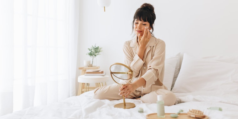 Woman doing self care applying beauty cosmetic, looking into mirror, while sitting in her bed