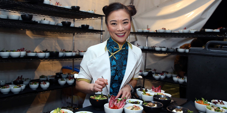 Shirley Chung smiles in a white jacket and blue Chinese-inspired blouse holding a fork over bowls of food