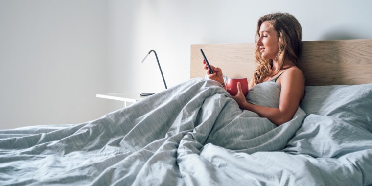 Woman sitting up in bed looking at smartphone, holding a red mug, wrapped in a grey duvet