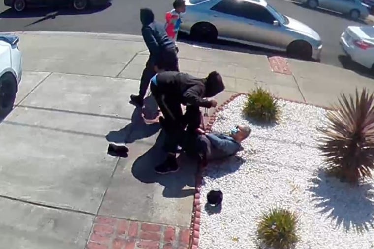 Still of surveillance camera footage showing one person in a black hoodie standing over an elderly Asian man who is on the ground and another person in a black hoodie facing away from them.