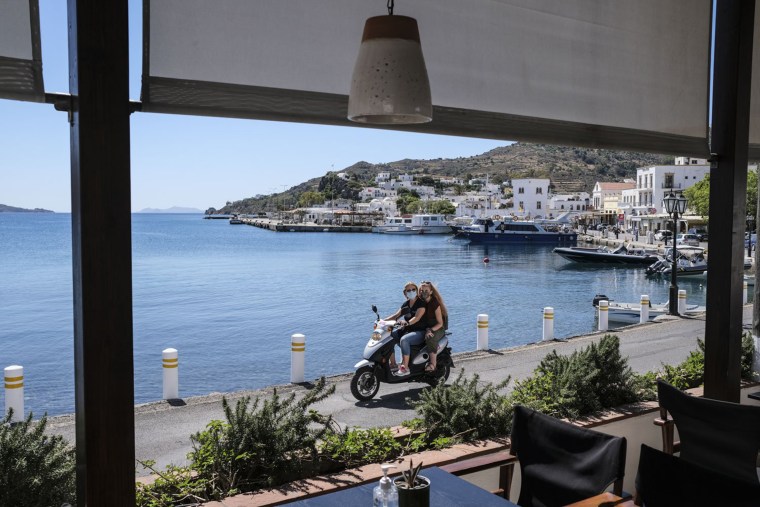 Image: People drive past a local cafe in the main port area of Patmos, Greece.
