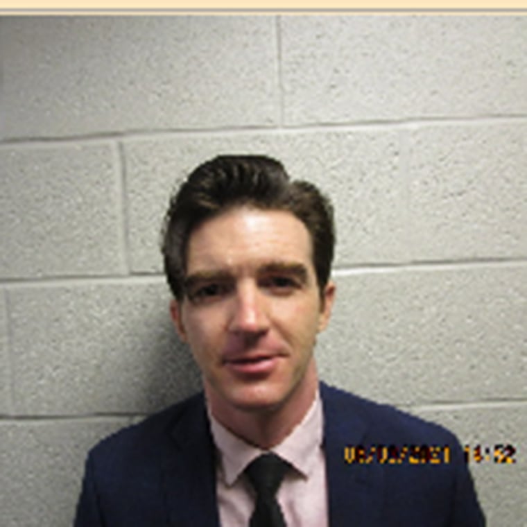 Drake Bell in a blue suit looks at the camera for a mug shot with a slight smirk on his face