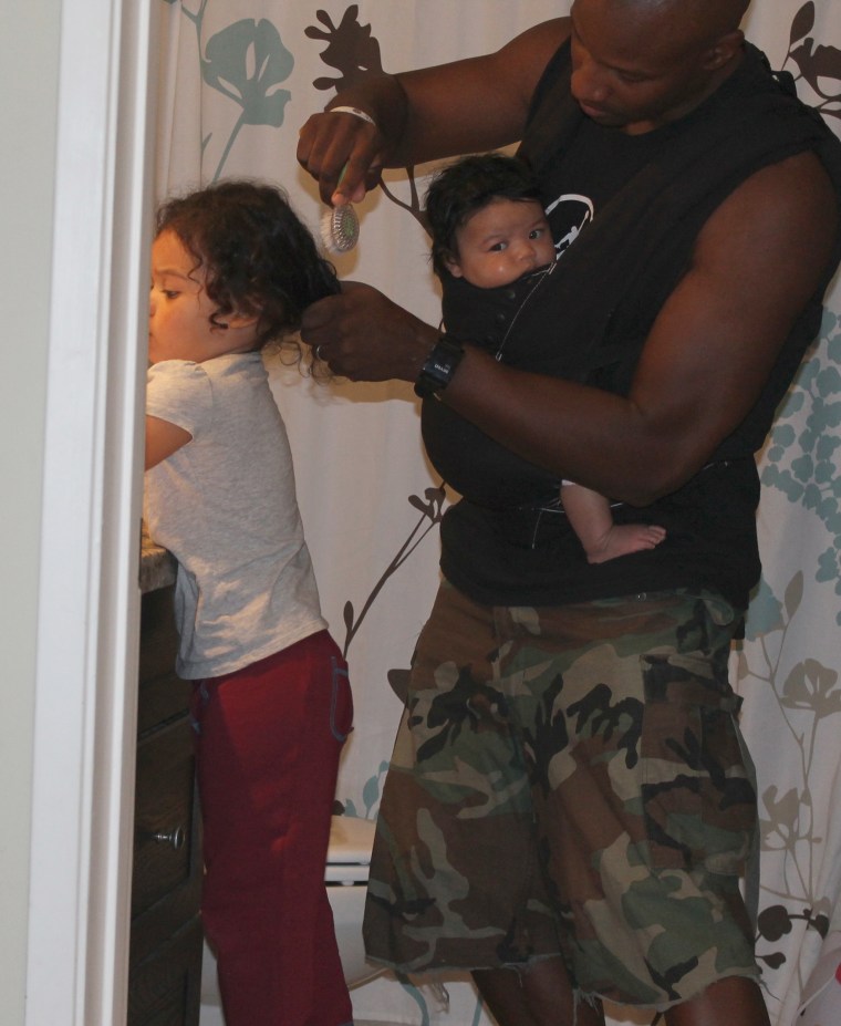 Doyin Richards does his older daughter's hair while wearing his baby girl in a baby carrier.