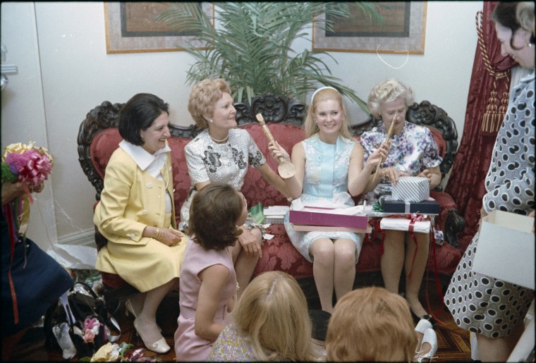 Tricia Nixon opening gifts at a bridal shower in her honor, hosted by the women of the press. First lady Pat Nixon, reporter Helen Thomas, and mother of the groom, Anne Cox, share the couch with her on May 26, 1971.