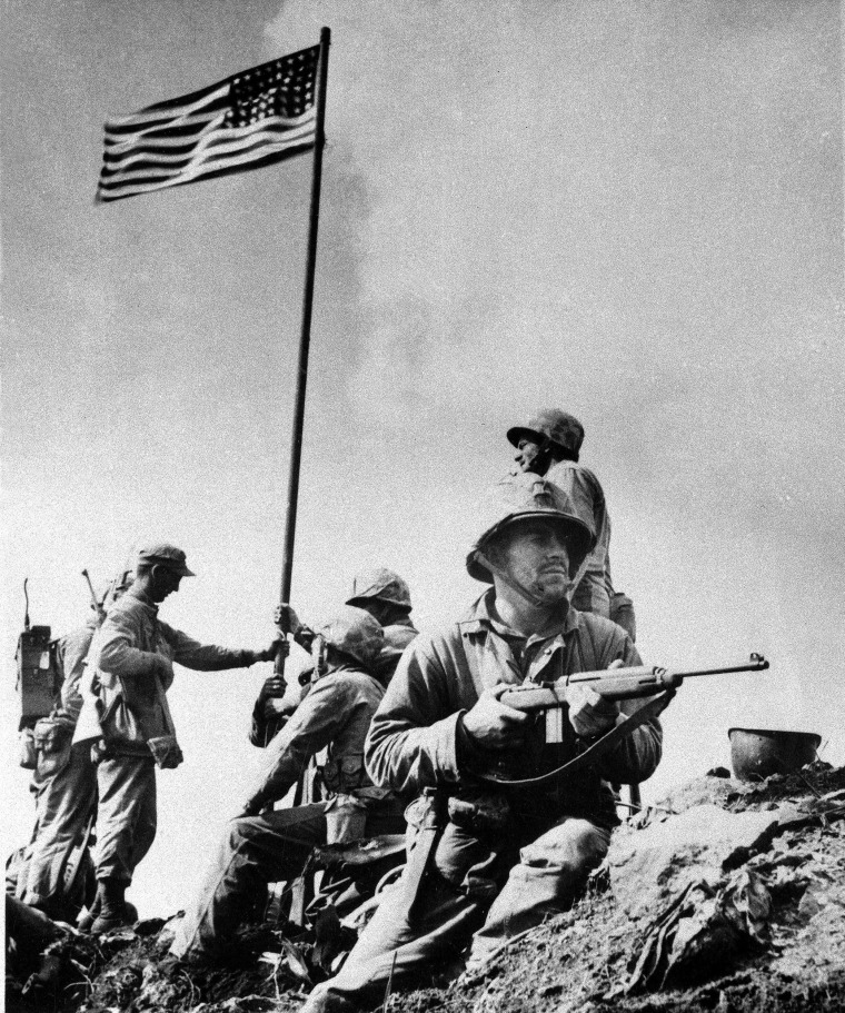 A U.S. Marine from the 5th Division of the 28th Regiment stands guard atop Mt. Suribachi at Iwo Jima in the Volcano Islands of Japan as others hoist the American flag during World War II, Feb. 23, 1945.