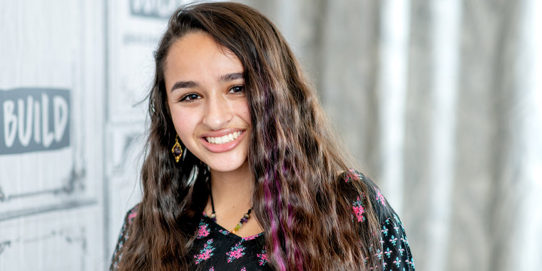 Jazz Jennings with a few streaks of purple in her hair smiles is a close up photo with a white background