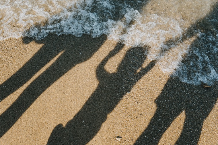 IMage: Shadows of a family holding hands on a beach as the ocean water rushes in.