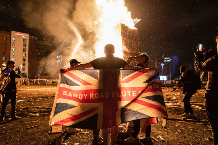 Image: A man drapes the Union Jack around his shoulders in front of a blazing pyre at the Sandy Row bonfire build in South Belfast.