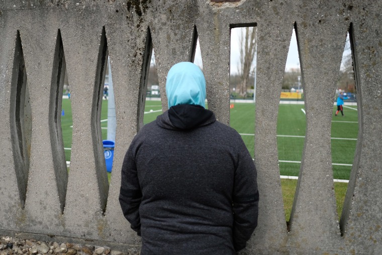 Image: A woman wearing a headscarf watches football training for children through a concrete fence in Leipzig, Germany