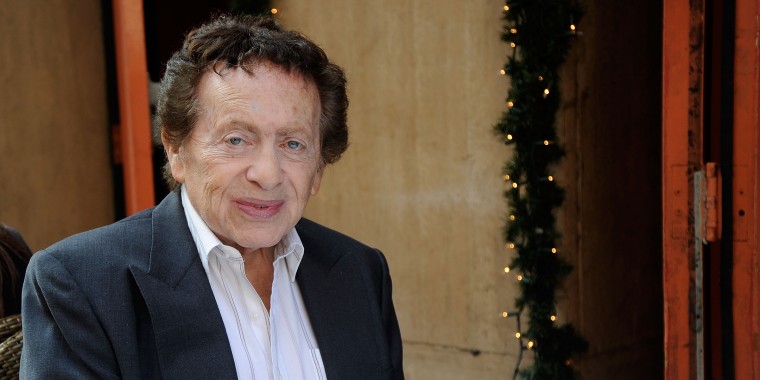 Jackie Mason sits in a suit at a Manhattan restaurant holding a credit card in front of Christmas decor