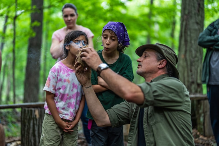 Shane Hobel puts an ember into cambium to start a fire while Sahira Pawria-Sanchez and Ara Bella Pajoohi blow on the ember.