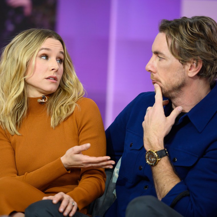 Kristen Bell gestures at her husband Dax Shepard while sitting on a TV set in front of a purple background