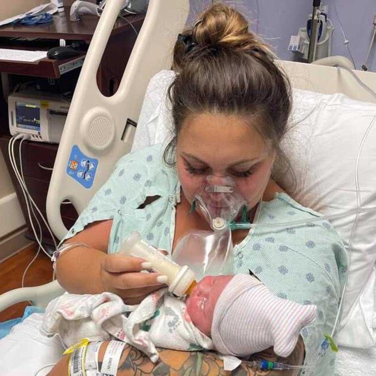 young woman still in hospital gown and bed holds newborn in her arms while wearing an oxygen mask
