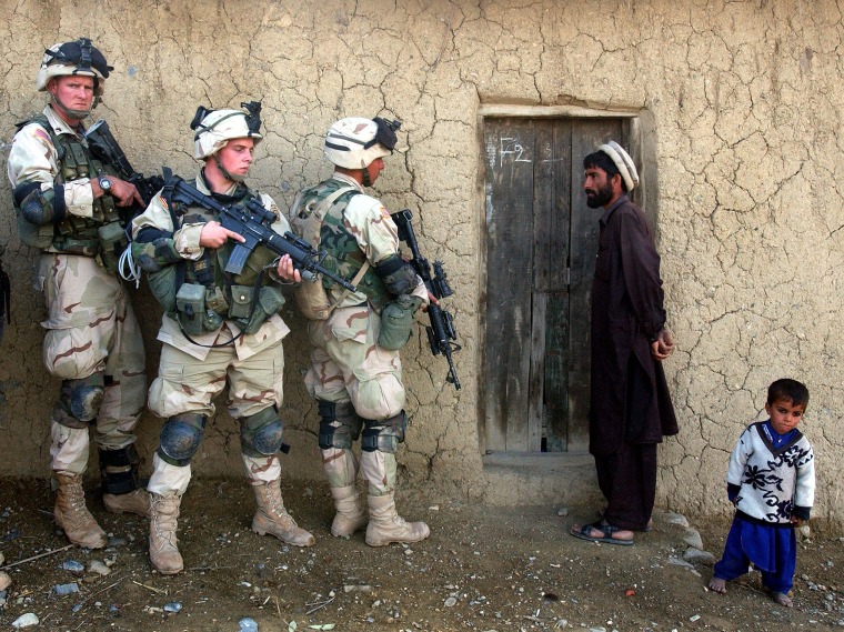 Image: An Afghan man and his son watch as soldiers from the U.S. Army 82nd Airborne Division prepare to sweep their home