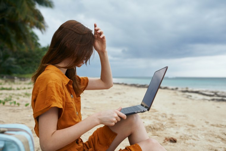 Woman sitting on the beach straightens her hair do not hold open working laptop on her lap.