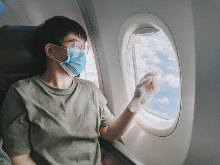 An asian chinese mid adult woman traveling in the commercial airplane passanger seat looking outside window with facemask and rubber surgical glove