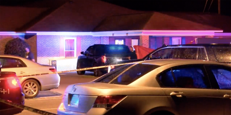 police lights illuminate a black parked SUV outside a low-rise brick building with yellow crime tape