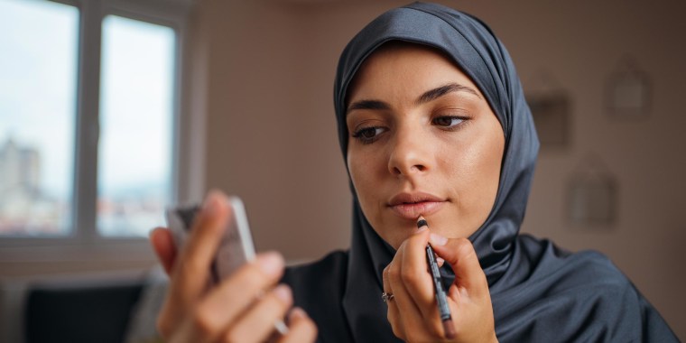 Young woman with hijab applying lip liner at home, while looking in handheld mirror