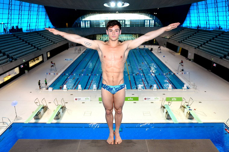 Tom Daley stands on the diving platform with his back to the water and both hands raised to his sides