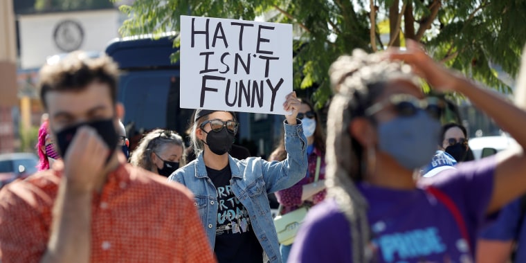 A person in a jean jacket and face mask holds a sign that reads "Hate isn't funny."