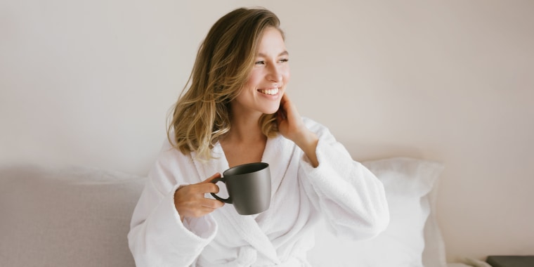 Woman wearing white bathrobe in bed holding a black mug