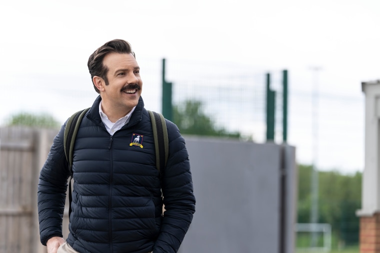 A mustachioed man in a navy puffer coat cheerfully walks through dreary london wearing a backpack