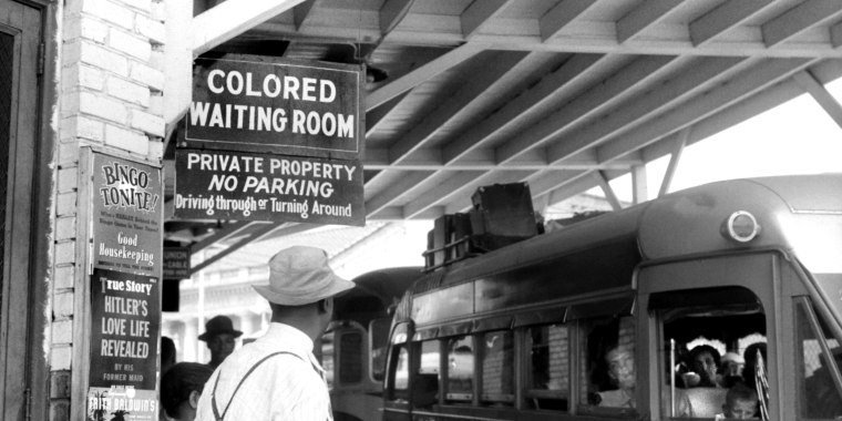 Image: People waiting at a bus station which has a sign that reads,\"Colored Waiting Room\".